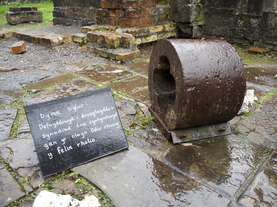 A small, rusted circular bearing next to a black sign with handwritten Welsh text.