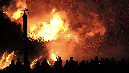 Photo of orange and yellow flames, with people and a forest in silhouette against the fire.