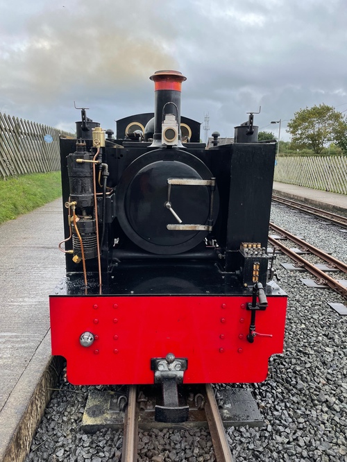 The very front of the black engine, with the face plate clearly visible. It has silver hands pointing to 5 o’clock, and a headlamp in front of the funnel.