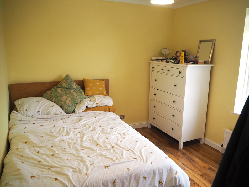 Looking towards one end of the bedroom. On the left hand side is a double bed pushed into one corner, with a white duvet cover with small cheetahs on it, plus a large stack of pillows. On the right hand side is a tall, white set of drawers, with a variety of small objects on top (mostly toys and books). The walls are painted a light yellow colour.