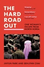 The cover of “The Hard Road Out”. Most of the cover is text, and then there’s a photo. A woman stands in a crowd of officials in brown uniform (presumably North Korean military attire). She has her back to the camera, and she’s wearing a baby pink dress that makes her stand out from the men milling around her.