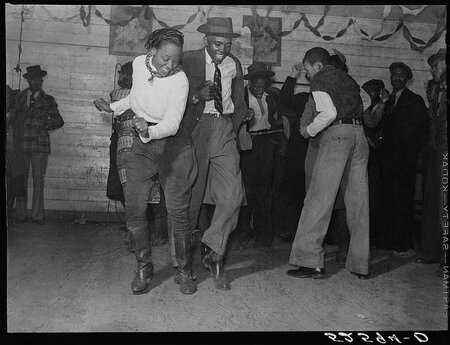 Black-and-white photo of a couple dancing side-by-side, with other people around them dancing or watching from the sidelines.