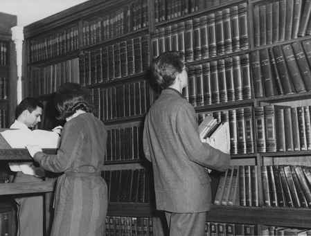 Black-and-white photo of people looking at a shelf of encyclopedias in a library. The man closest the camera is already holding a stack of papers, and looking upwards owards one of the books.