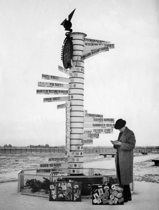 Black-and-white photo of a signpost with arrows pointing in all different directions, with a man looking down at a map and pondering his next move.