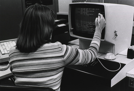 A person with long, dark hair using a computer terminal. They're facing away from the camera, and holding some sort of stylus up to a monitor showing monospaced text.