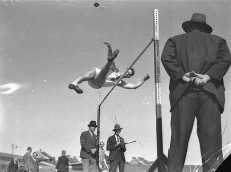 Black-and-white photo of a man passing over a high jump bar, as two officials watch from the sideline.