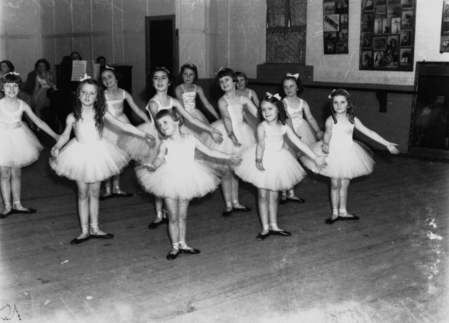 A black-and-white photo of young girls in white tutus, their arms held out in ballet poses, inside a dance studio.