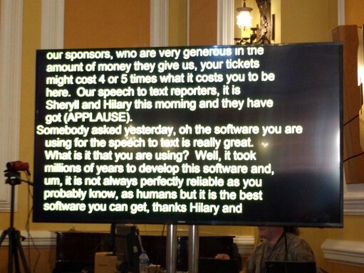 A television screen with yellow text on a black background, showing a transcript of somebody speaking.