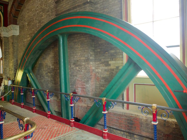 A large green wheel, with the top half visible, set against a brown brick wall and a window.