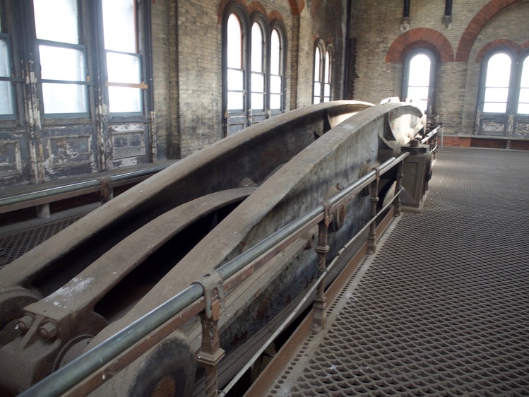A large, rusting metal pumping beam. It's against a wall, rising in the background, with windows in the walls.