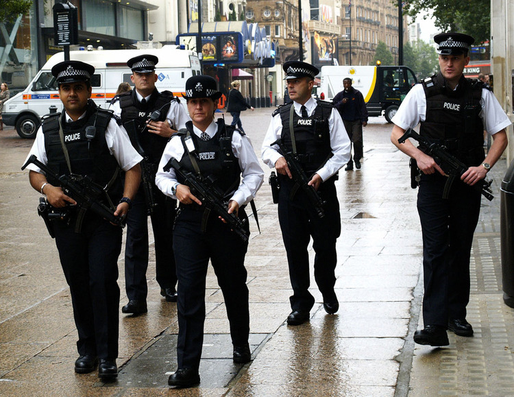 Five police officers walking down a street, all holding large guns which are pointed towards the ground.