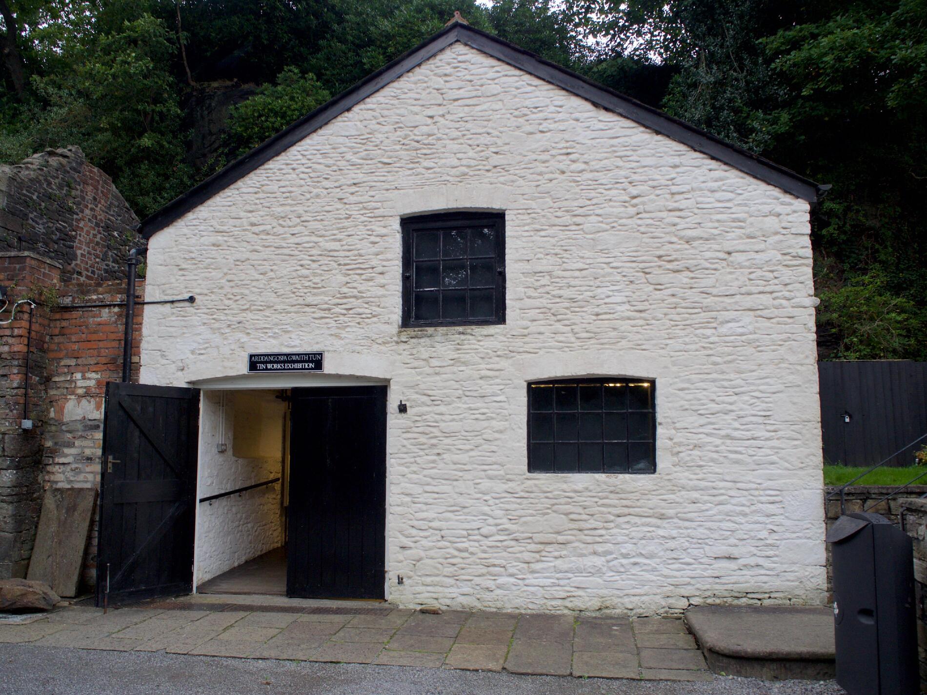 A white building with two windows, black doors, and a small sign reading “Tin Works exhibition”.