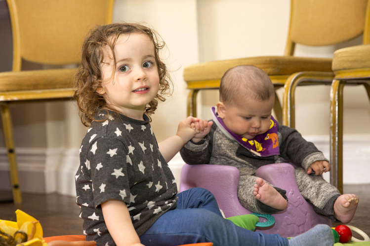 Two babies sitting on the floor with some chairs and toys.