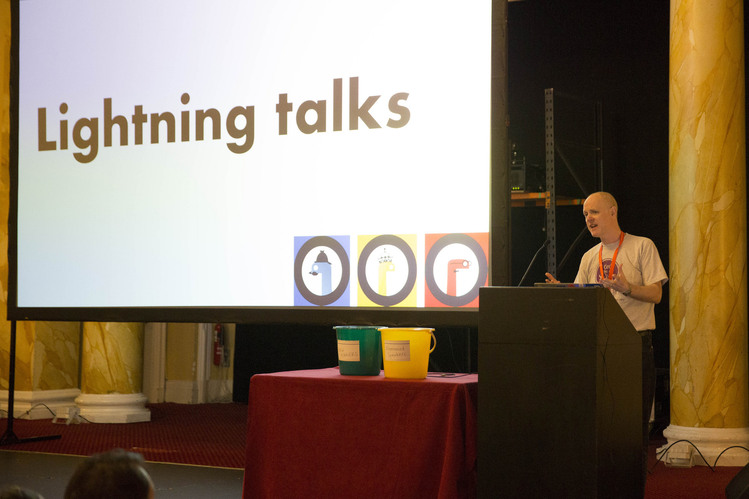 A person at a lectern with a slide behind them saying “Lightning talks” and two buckets on the table.