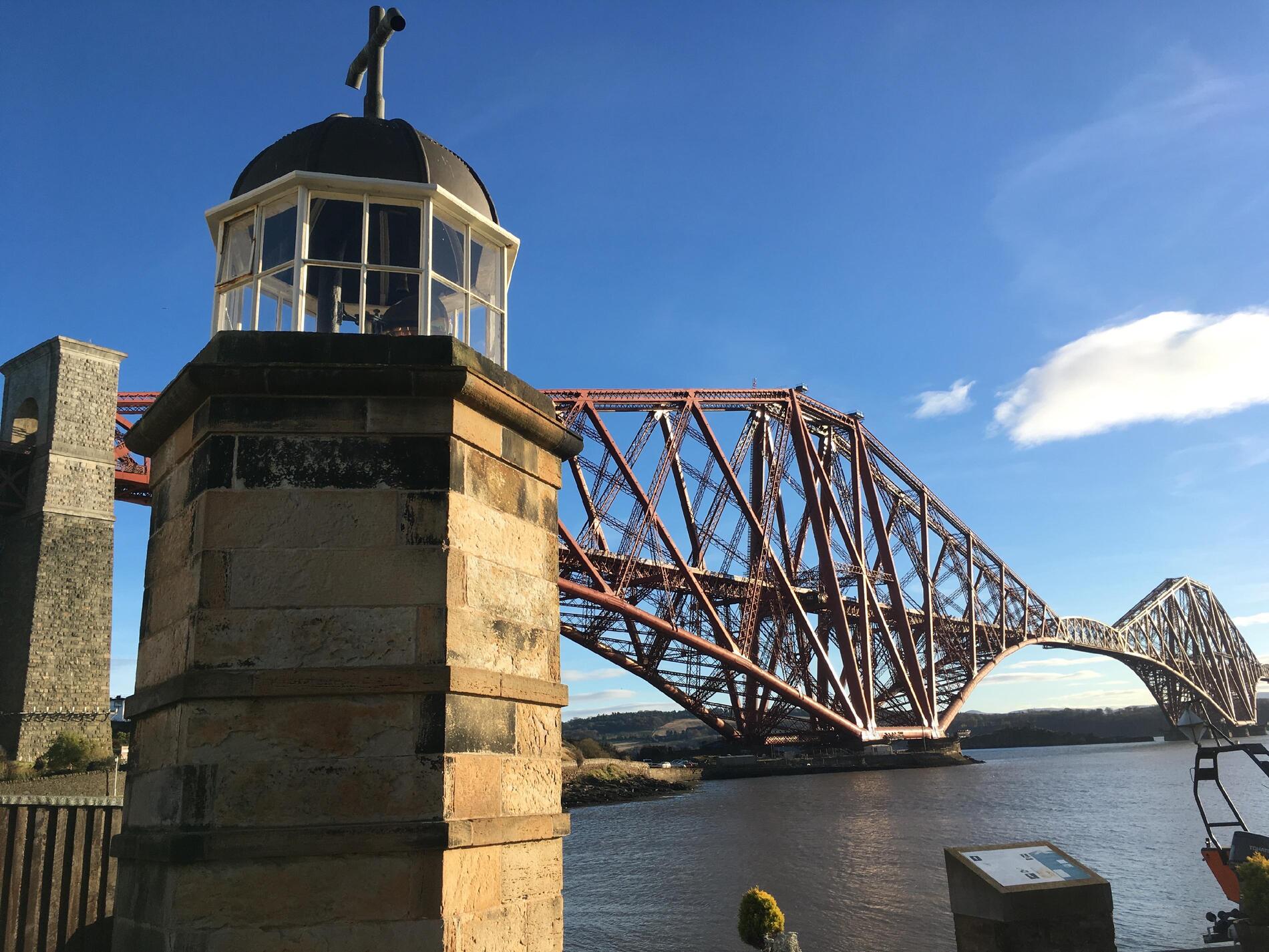 The lighthouse in the foreground on the left, with the bridge set against a blue sky in the background.