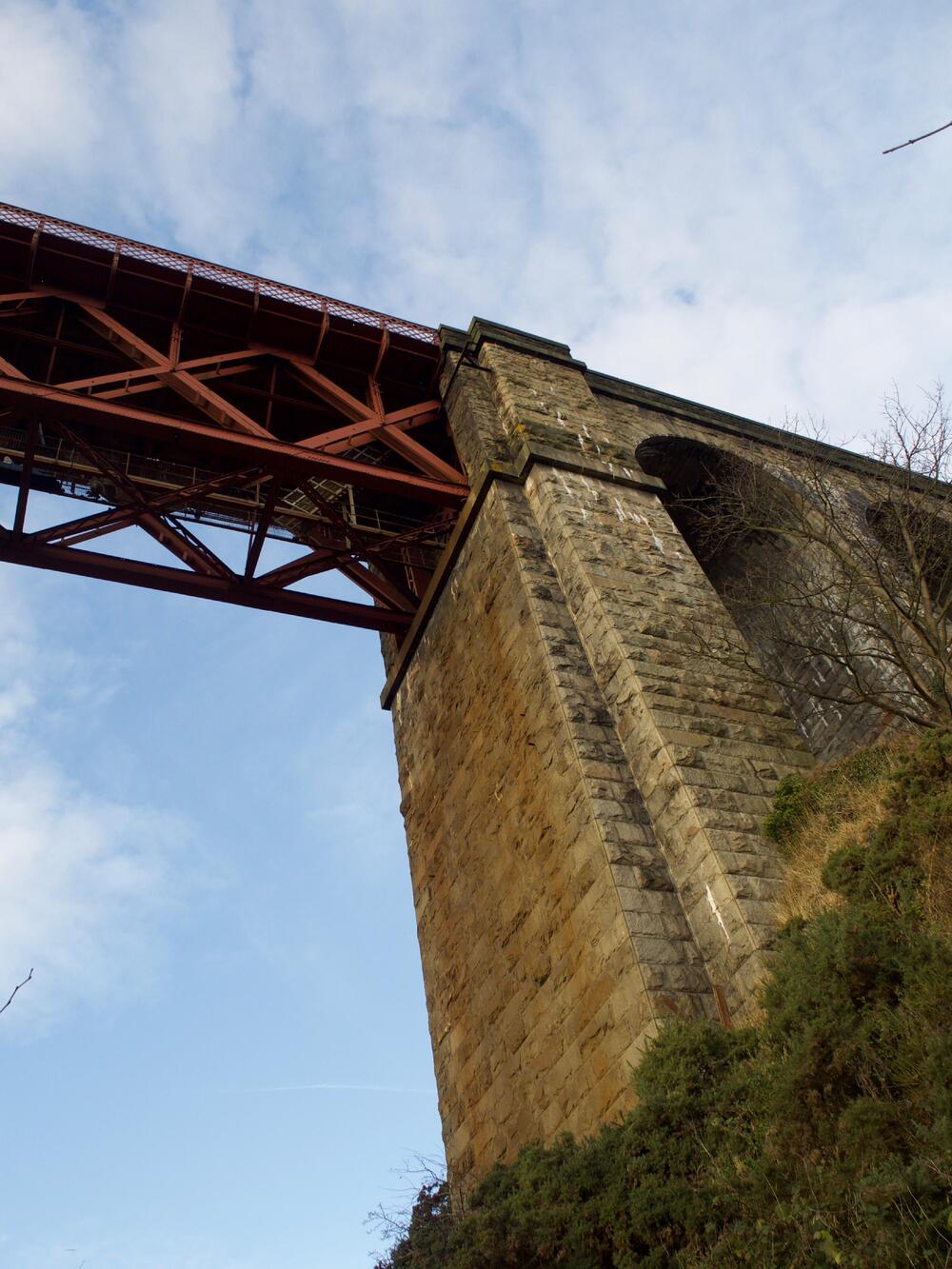 A yellowish-coloured stone viaduct, with the red girders of the bridge atop it.