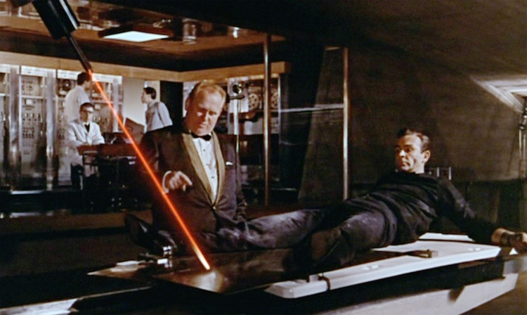 A man in all black (James Bond) is strapped to a table as another man in a golden suit (Goldfinger) overlooks. A large red laser is cutting through the table.