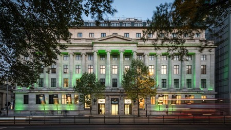 Photograph of the front elevation of the Wellcome Collection building at dusk. The building is illuminated with a series of up-lighters, flooding the facade with green light. In the foreground at ground level are blurred streaks of red and yellow from the headlights and tail lights of passing vehicles. The leaves of trees to the left and right of the image frame the building and are themselves blurred as a result of the wind and long photographic exposure.