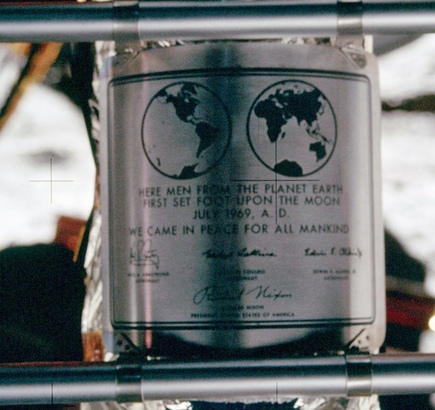A metal plaque attached to a larger structure. It has two hemispheres of the Earth, followed by some text (below), and signatures of the three astronauts and the then-US president.