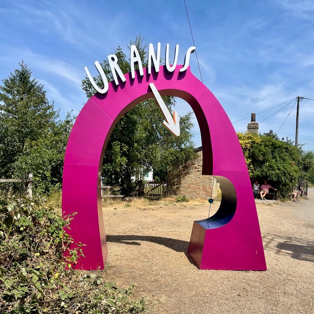 A magenta arch labelled 'Uranus', sitting on a dried-out patch of grass. There are a couple of small buildings in the background, which are part of a river lock mechanism that's out of frame.