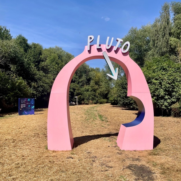 A baby pink archway labelled 'Pluto', in an open area filled with dried-out grass. There are a few tufts of green grass, but mostly it's all straw.