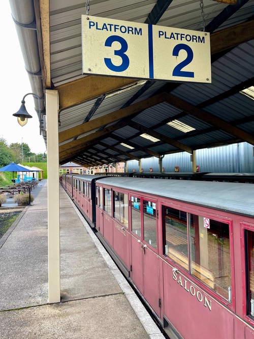 Looking down a train platform with two overhead signs for “Platform 3” and “Platform 2”. Sitting below the sign in platform 2 are a row of burgundy carriages. The carriages are about a metre high, with windows running along the top half, and split into doors for the different compartments. Some of the doors are labelled – for example, the closest door is labelled “Saloon”.