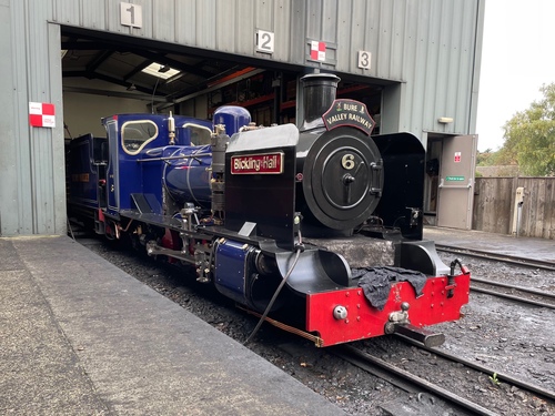 A dark blue steam engine, sitting in the sidings. It has a burgundy nameplate “Blickling Hall”, the number “6”, and a head plate “Bure Valley Railway”. It’s sitting partway out of a large grey, box-shaped shed. Partially visible is a tender inside the shed – this is a tender engine. Like the black engine, this has red plates on the wheels and gold trim.