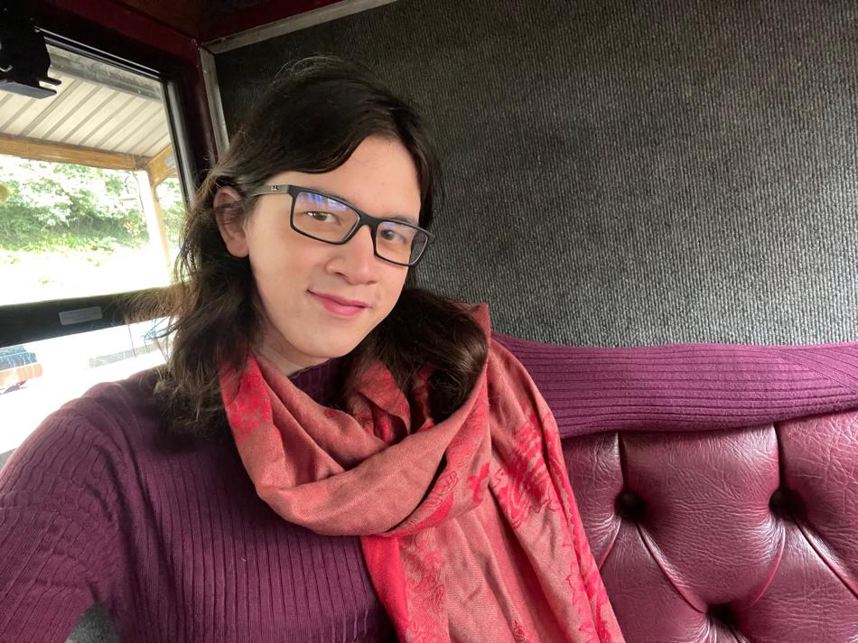 A selfie! I’m sitting on one of the seats in a maroon-coloured jumper and a red scarf, with my right arm outstretched over the top of the seat. I’m wearing glasses, dark-coloured lipstick, and I have dark brown hair falling on either side of my face. I’m tilting my head a bit, and smiling – because my jumper is the same colour as the seat! (Well, almost.)