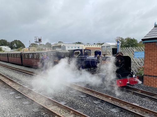 A dark blue tender engine, blowing copious clouds of white steam from the very front of the engine. Behind it are several low-height coaches, which it’s just pulled along half the journey.