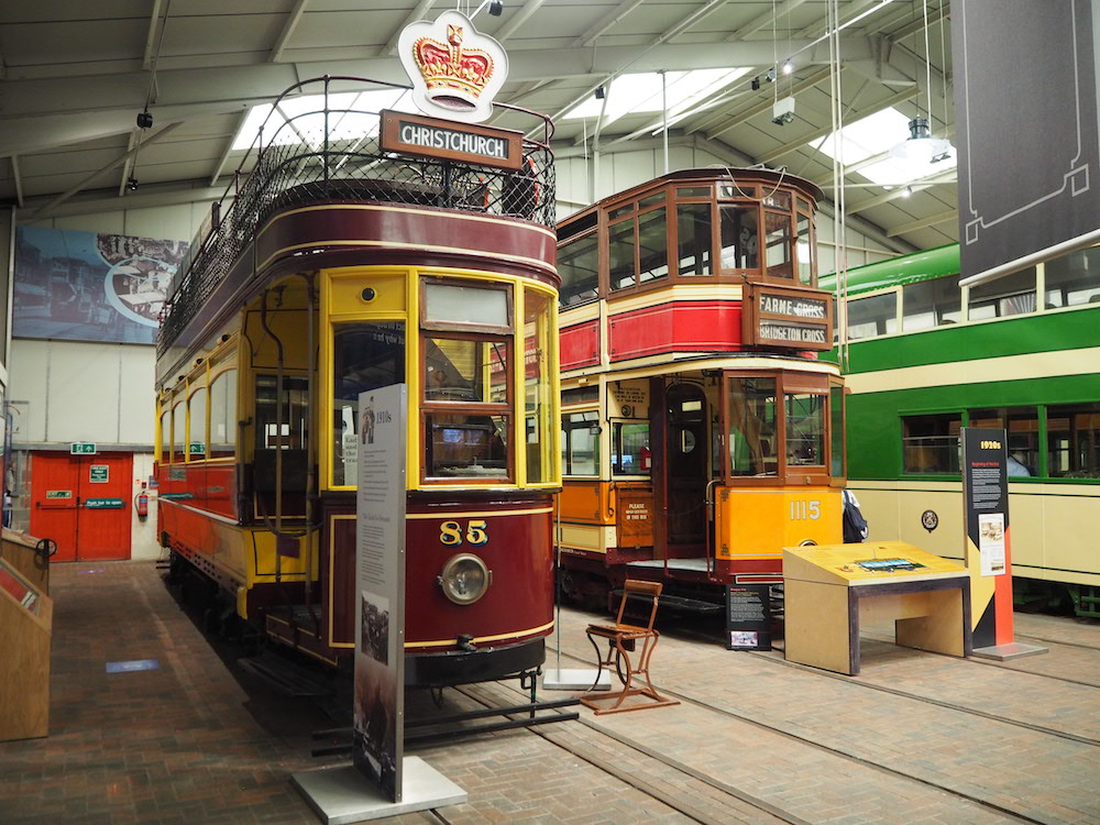 Two double-decker trams standing on tracks in a gallery space. The left-hand tram has a dark red and yellow livery, with an open-air upper deck, the number '85' and a destination board 'Christchurch'. The right-hand tram has an orange, red and cream livery, and the departure board 'Farme Cross / Bridgeton Cross'.