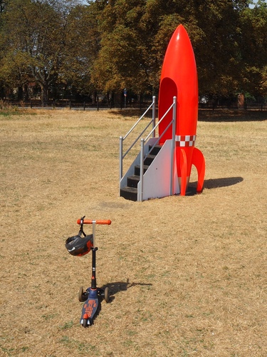 A red rocket with steps leading into the back of it, where kids can climb up and have their photo taken through the porthole on the other side. There's also an abandoned scooter standing near the rocket, whose child has apparently wandered off.