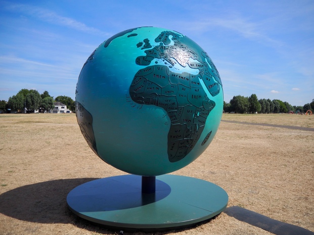 A large turquoise and teal globe, showing the Earth. There's a thin border around each country, and in each country is the word 'us' (in blue) and 'them' (in red). The globe is turned so that Africa is closest to the viewer, with part of Europe visible in the top.