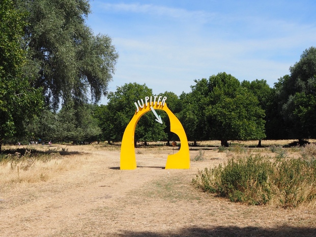 A bright yellow arch labelled 'Jupiter' and an arrow pointing to the planet. The planet model is visible from quite a distance away. The arch is in a large park with parched dry grass and some green trees.