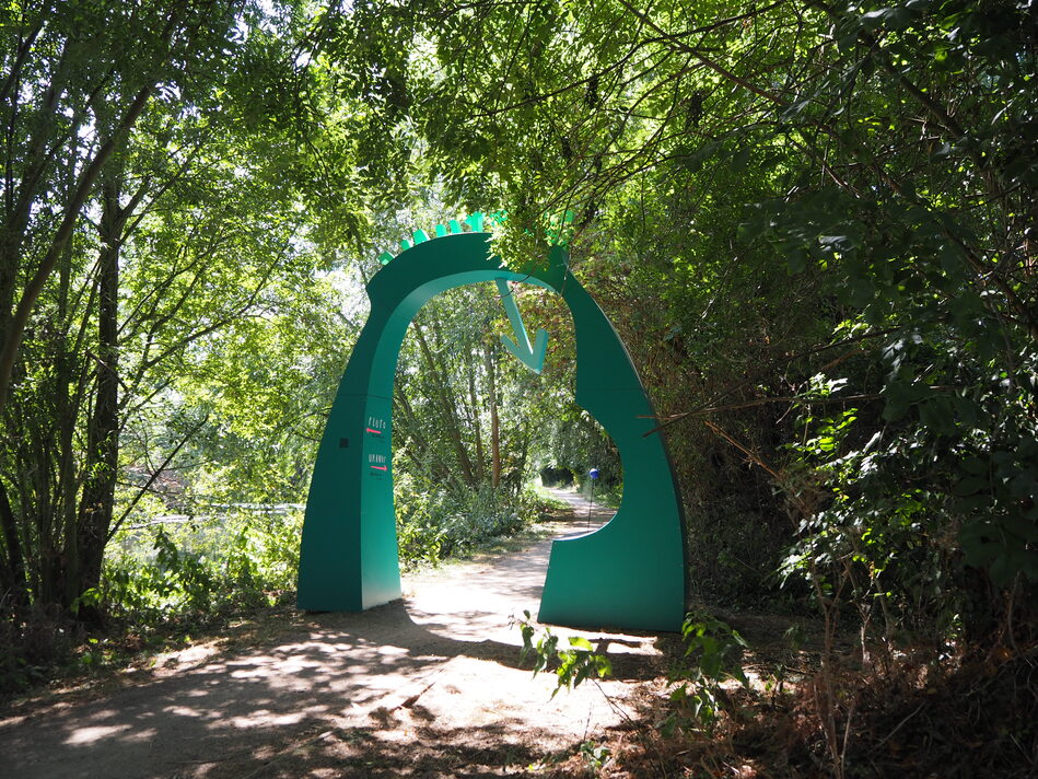 The back of a green-ish arch for 'Neptune', looking back along the trail we'd just walked. The arch is surrounded by trees, which cast interesting shadows on the ground and break up the profile. The path curves to the left in the distance, and you can just see the river through the trees.