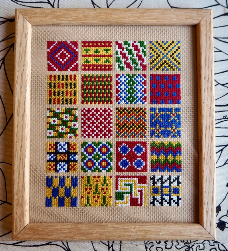 A cross-stitch piece embroided on a brown fabric, with twenty brightly-coloured squares laid out in a grid. Each square uses a couple of colours (red, blue, green, yellow, white, or black) and has a simple repeating pattern. For example, one square is yellow with repeating black and red vertical lines; another has a red background with concentric white diamonds.