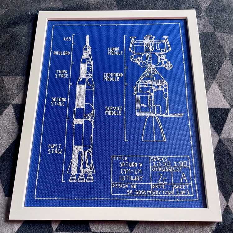 A picture in a white frame. It looks a bit like a blueprint, with white thread on a blue background. On the left hand-side is a tall, sleek rocket (the Saturn V) and on the right are the various stages of the Apollo lander (lunar module, command module, and service module). In the lower right-hand corner is some technical information, including a title and a scale.