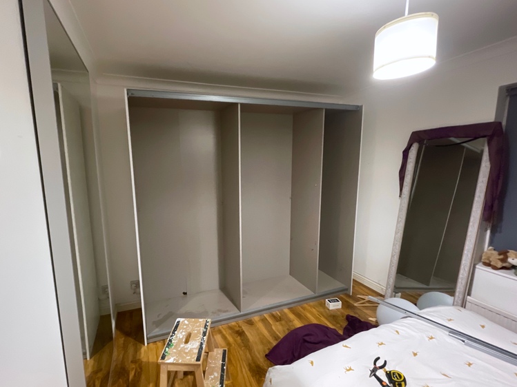 The same bedroom from a previous angle, with a partially disassembled wardrobe against one wall. The doors have been removed and you can see the empty interior, which shows three vertical compartments separated by dividers. The interior is a drab and uninspiring grey, with dark water stains in several parts.