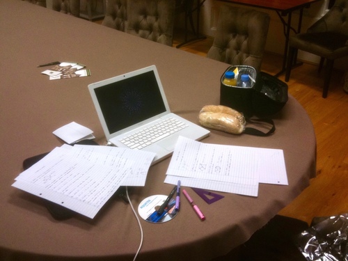 A white laptop on a brown table, surrounded by handwritten papers and a half-finished lunchbox. A couple of diagrams are visible on the papers, drawn in purple ink.