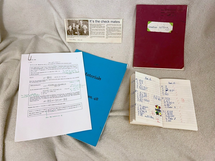 A collection of a few papers, including a purple exercise book, a blue workbook, a small book with some handwritten numbers, and a newspaper clipping about a chess club.