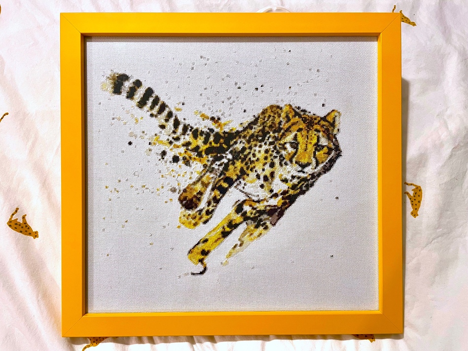 Some cross-stitch embroidery of a cheetah. The cheetah is running, with it legs angled beneath it as if its partway off the ground. It's leaning towards the right, with its tail curving behind it. The piece is stitched on white fabric in varying shades of yellow/brown/black, and mounted in a square frame made of yellowy-orange wood.