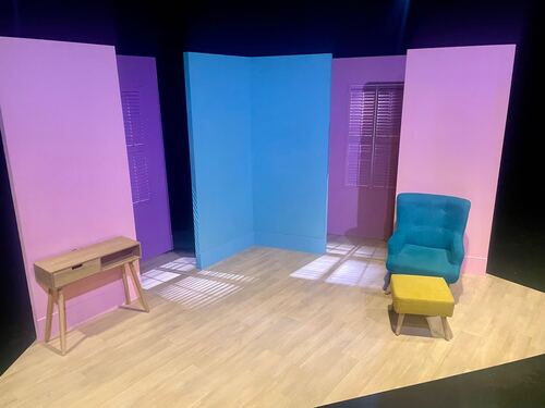 An empty theatre set. There are plain walls in pink and purple, with a small side table on stage right and a comfy chair on stage left. Otherwise, the set is empty.