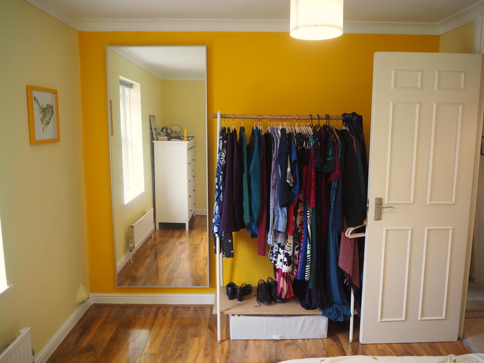 Looking towards the other end of the room, with the end of the bed just visible in the bottom right-hand corner. The wall is a bright, egg-yolk yellow. On the left-hand side is a mirror that's almost the full height of the wall. To the right of the mirror is a clothes rail with various clothes hanging from it, and a few pairs of shoes on the floor below. To the right of the clothes rail is the open bedroom door, hiding the corner behind it.