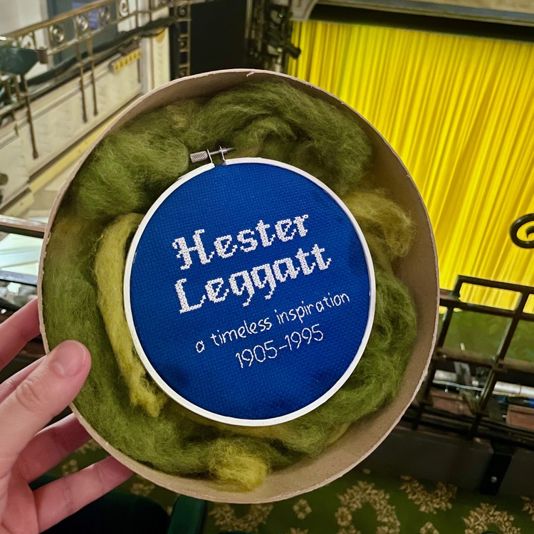 Some embroidery on blue fabric with a circular frame. There's white text that says “Hester Leggatt / a timeless inspiration / 1905–1995”. I’m holding it in the auditorium of the Fortune Theatre, where the play is being shown – some seats are visible in the background, along with the bright yellow curtain on stage.