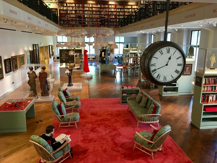 The Reading Room at Wellcome Collection. A large, brightly-lit room with shelves interspersed with museum objects and comfy chairs.