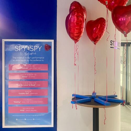 A blue poster board titled “Spy for Spy” with a message: “The order of today’s performance as determined by the audience is:” followed by six pink cards, each with a title and a song name. Next to the poster are several shiny red heart balloons.