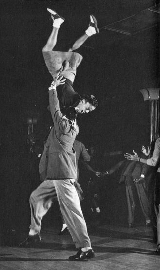 Black-and-white photo of two dancers. The leader has lifted their follower up in the air, so the follower is upside down, their head facing the floor and her feet kicking towards the ceiling. She seems to be crying in delight (I hope!).