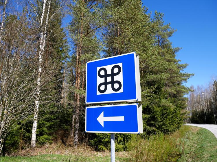 A large road sign in a forest, with a black symbol that looks like a square with a loop on each corner.