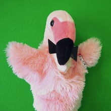 A small flamingo hand-puppet with a black bow tie on its neck, with a flamingo neck and two fluffy, flappy arms.