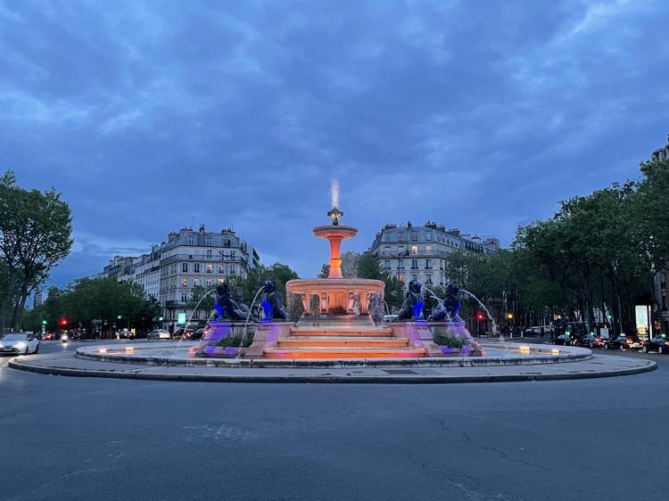 A water fountain in the middle of a roundabout, lit up in orange and purple lighting from below. Around the centre of the fountain are four creatures on pedestals (cats?) with water coming out of their mouths.