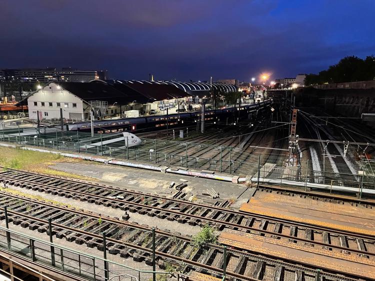 A nighttime photo of a large rail yard with many parallel and overlapping tracks. A long passenger train can be seen in the background of the shot, largely visible from the light coming from inside the windows.